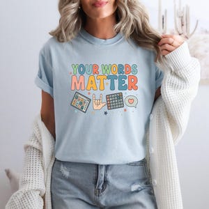 May include: Light blue t-shirt with the text "YOUR WORDS MATTER" in colorful, retro-style lettering. The shirt features illustrations of a tablet, a hand signing "I love you" in ASL, and a calculator. A heart and speech bubble are also included.