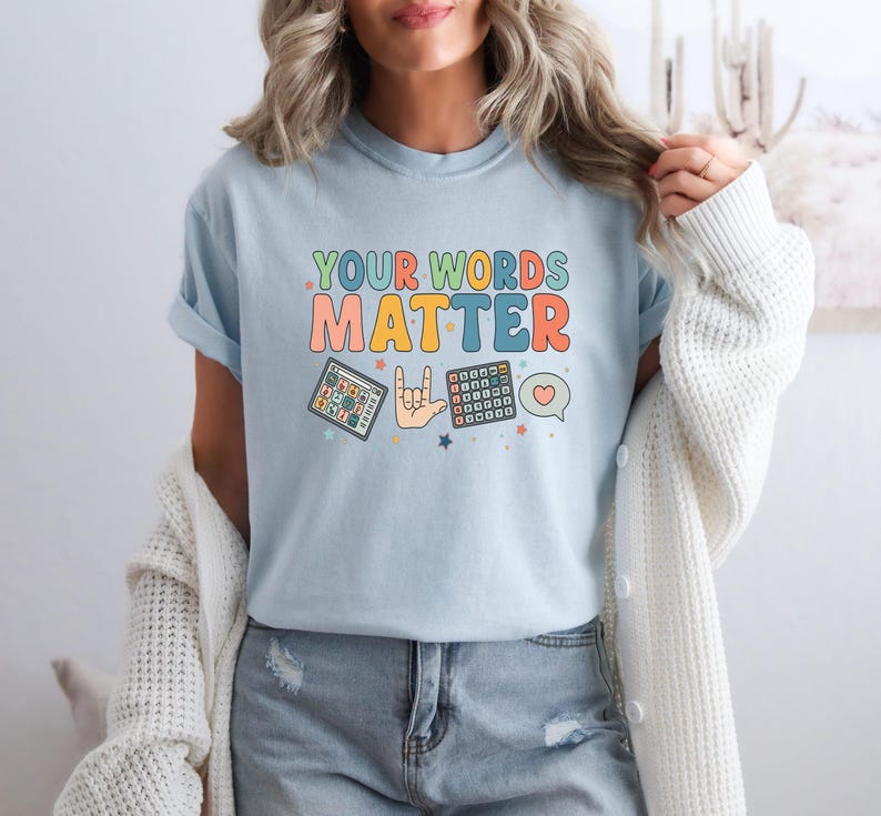 May include: Light blue t-shirt with the text "YOUR WORDS MATTER" in colorful, retro-style lettering. The shirt features illustrations of a tablet, a hand signing "I love you" in ASL, and a calculator. A heart and speech bubble are also included.