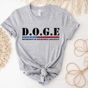 May include: A light gray t-shirt with the text "D.O.G.E" in bold black letters, above a red and blue striped design. Below the stripes, the text reads "DEPARTMENT OF GOVERNMENT EFFICIENCY" in black.