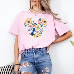 Op de afbeelding: Een roze T-shirt met een bloemig Mickey Mouse-ontwerp. Het ontwerp bevat gele, roze, blauwe en groene bloemen.