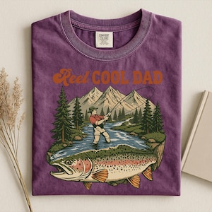 May include: Purple t-shirt with a graphic of a fisherman, mountains, and a large fish. The text "Reel Cool Dad" is in orange script. The shirt is folded, with a tag visible at the top.