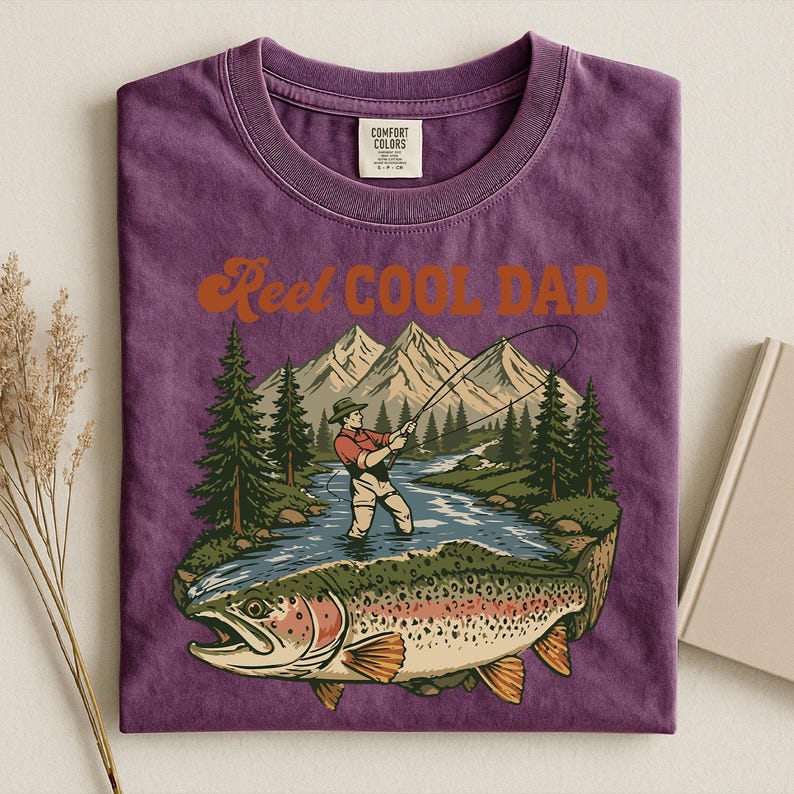 May include: Purple t-shirt with a graphic of a fisherman, mountains, and a large fish. The text "Reel Cool Dad" is in orange script. The shirt is folded, with a tag visible at the top.