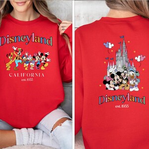Puede incluir: Sudadera roja con un logotipo de Disneyland colorido y un gr&aacute;fico de Mickey Mouse y sus amigos frente al castillo de Disneyland. El texto en la sudadera dice "Disneyland California est. 1955".