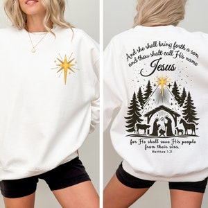 May include: White sweatshirt with a gold star design on the front and a Nativity scene on the back. The back design includes text that reads "And she shall bring forth a son, and thou shalt call His name Jesus for He shall save His people from their sins. Matthew 1:21."