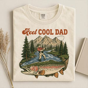 May include: Cream-colored t-shirt with a graphic featuring a fisherman in a river, mountains, and a large trout. The text "Reel Cool Dad" is in a retro font. The shirt has a Comfort Colors tag.