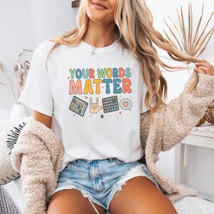 May include: White t-shirt with the text "YOUR WORDS MATTER" in colorful retro font. The shirt features illustrations of a tablet, a hand making a rock on gesture, a keyboard, and a heart-shaped speech bubble.