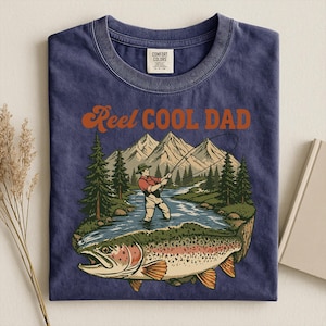 May include: Navy blue t-shirt with a graphic of a fisherman in a river, mountains, and a large trout. The text "Reel Cool Dad" is in orange script. The shirt is made of Comfort Colors material.