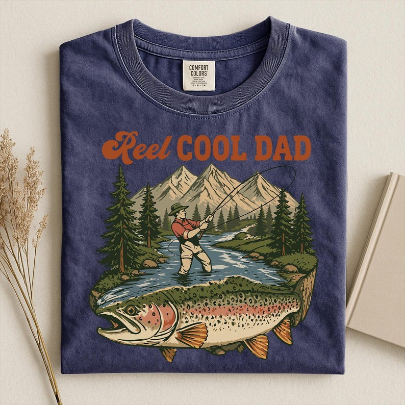 May include: Navy blue t-shirt with a graphic of a fisherman in a river, mountains, and a large trout. The text "Reel Cool Dad" is in orange script. The shirt is made of Comfort Colors material.