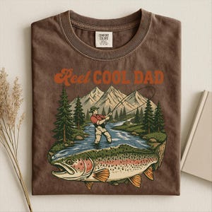 May include: Brown t-shirt with the text "Reel Cool Dad" in orange. The graphic features a fisherman in a river with a large trout, mountains, and trees. The shirt is made by Comfort Colors.