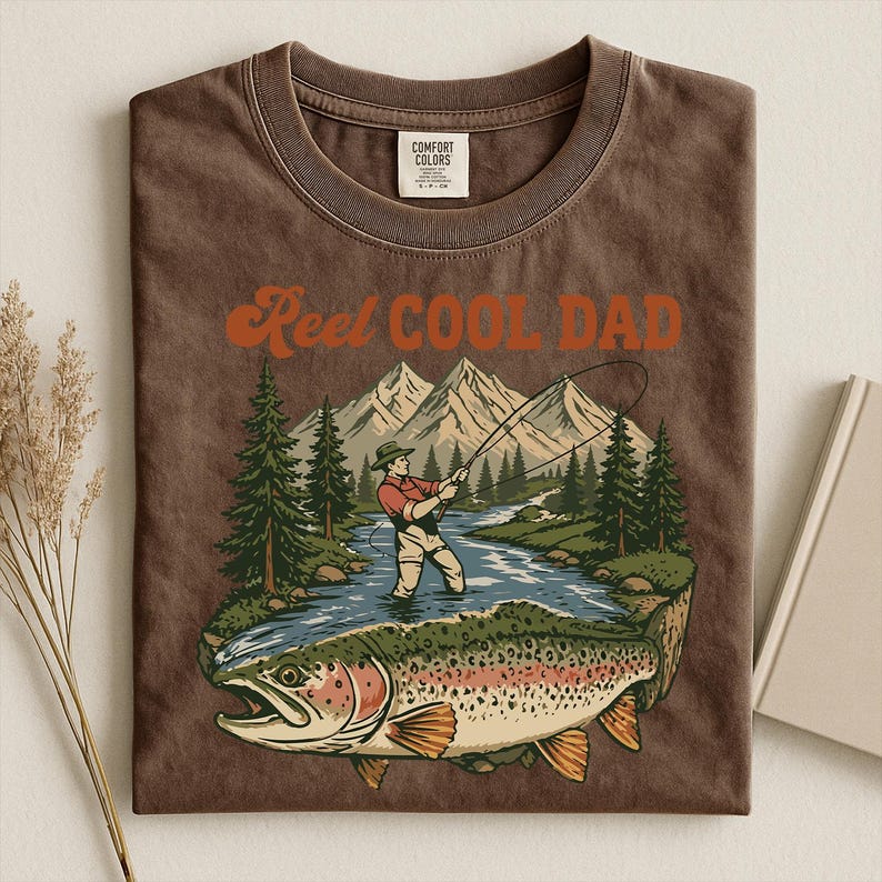 May include: Brown t-shirt with the text "Reel Cool Dad" in orange. The graphic features a fisherman in a river with a large trout, mountains, and trees. The shirt is made by Comfort Colors.