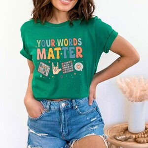 May include: A green t-shirt with the text "YOUR WORDS MATTER" in colorful letters. The shirt features graphic illustrations of a calculator, a hand signing "I love you" in sign language, and a speech bubble with a heart.