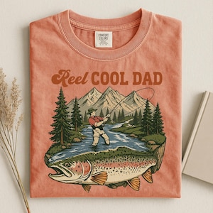 May include: Coral-colored t-shirt with a graphic of a fisherman in a river, mountains, and a large fish. Text reads "Reel Cool Dad" in brown script. The shirt has a classic crew neck and a tag at the top.