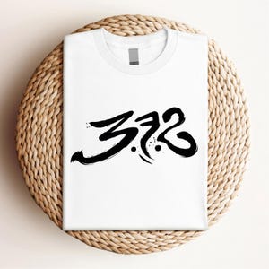 May include: White t-shirt with the black text "372" in a stylized, brushstroke font. The shirt is displayed on a round, woven wicker placemat.