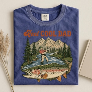 May include: Blue t-shirt with a graphic of a fisherman fly fishing in a river, with a large trout in the foreground. The text "Reel Cool Dad" is above the graphic in orange lettering. The shirt is folded.