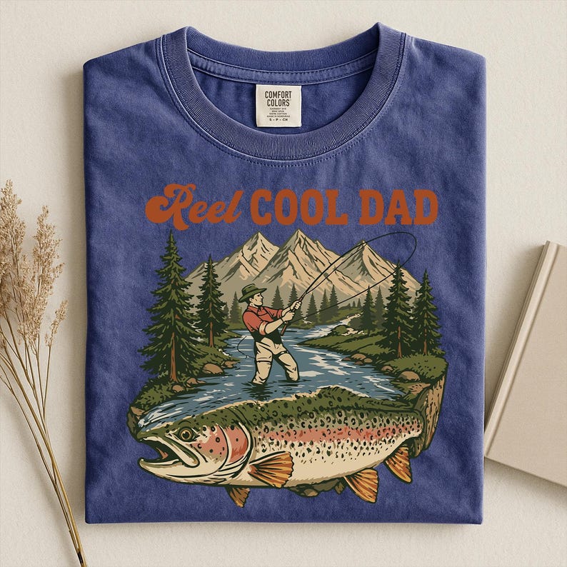 May include: Blue t-shirt with a graphic of a fisherman fly fishing in a river, with a large trout in the foreground. The text "Reel Cool Dad" is above the graphic in orange lettering. The shirt is folded.
