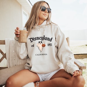May include: Beige hoodie with the text "Disneyland est 1955 CALIFORNIA". The graphic features a ghost with Mickey Mouse ears holding a jack-o'-lantern. The hoodie has a drawstring hood and a front pocket.