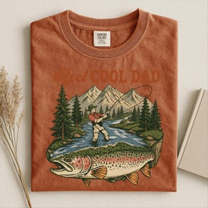 May include: Burnt orange t-shirt with a graphic of a fisherman fishing in a river, with a large trout in the foreground. The text "Reel Cool Dad" is arched above the graphic. The shirt is folded.