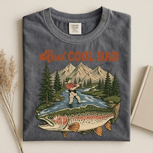 May include: Dark gray t-shirt with the text "Reel Cool Dad" in orange. The graphic features a fisherman in a river with a large trout, mountains, and trees. The shirt is a Comfort Colors brand.