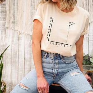 May include: Cream-colored t-shirt featuring a black graph with the coordinates (6,7). The graph has an x and y axis with numbers. The shirt is made of a soft material.