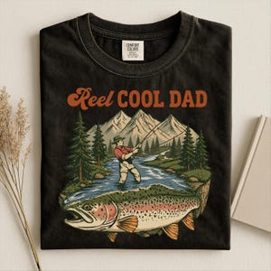 May include: Black t-shirt with a graphic of a fisherman in a river, mountains, and a large fish. The text "Reel Cool Dad" is in orange script. The shirt is made by Comfort Colors.