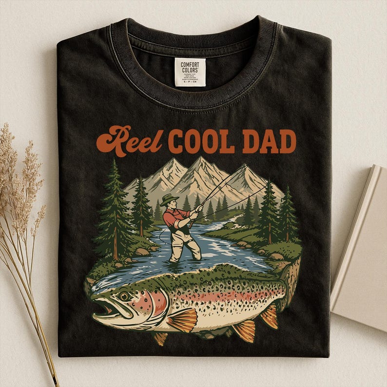 May include: Black t-shirt with a graphic of a fisherman in a river, mountains, and a large fish. The text "Reel Cool Dad" is in orange script. The shirt is made by Comfort Colors.