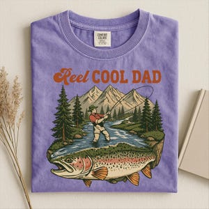 May include: A purple t-shirt with the words "Reel Cool Dad" in orange lettering. The graphic depicts a fisherman in a river with a large fish, mountains, and trees.