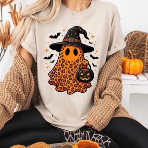 May include: A beige t-shirt featuring a graphic of an orange ghost with a black leopard print pattern. The ghost wears a black witch hat and carries a black jack-o'-lantern bucket. Black bats and orange stars surround the ghost on the shirt.