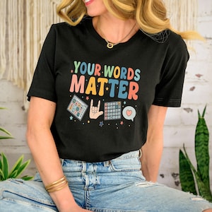 May include: Black t-shirt with the text "YOUR WORDS MATTER" in colorful letters. The shirt features graphic illustrations of a keyboard, a hand making a gesture, and a heart-shaped speech bubble. The shirt is worn with blue jeans.