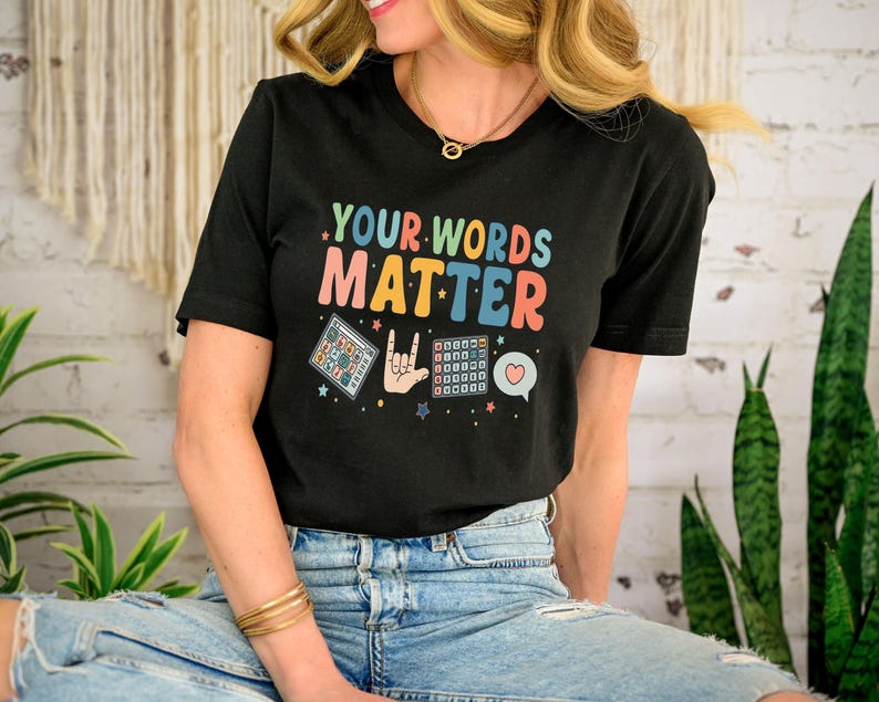 May include: Black t-shirt with the text "YOUR WORDS MATTER" in colorful letters. The shirt features graphic illustrations of a keyboard, a hand making a gesture, and a heart-shaped speech bubble. The shirt is worn with blue jeans.