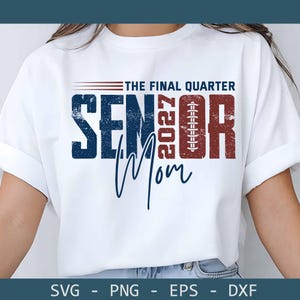 May include: A white short-sleeve t-shirt featuring a graphic with the text THE FINAL QUARTER SENIOR 2027 MOM. The design includes a football illustration replacing the letter I in SENIOR, with distressed blue and red lettering on the front.