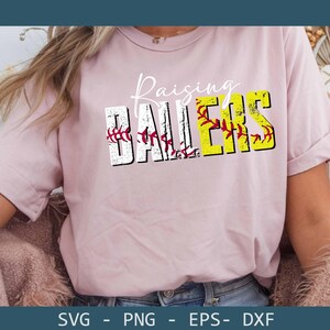 May include: A light pink t-shirt with the words "Raising Ballers" in a distressed font. The word "Ballers" is in yellow and white with baseball stitching details. The shirt is a casual style.