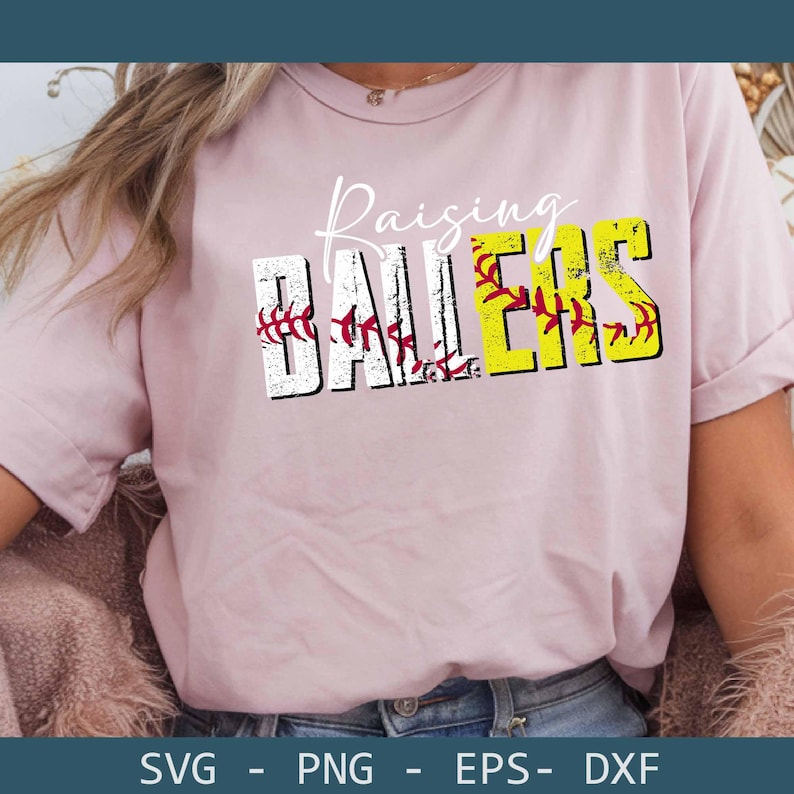 May include: A light pink t-shirt with the words "Raising Ballers" in a distressed font. The word "Ballers" is in yellow and white with baseball stitching details. The shirt is a casual style.