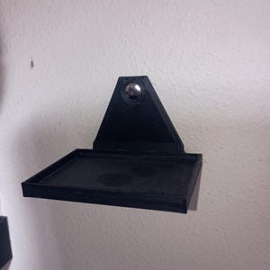May include: A black, wall-mounted shelf with a triangular top and a rectangular base. The shelf is attached to the wall with a single silver screw. The shelf is made of a matte black material.