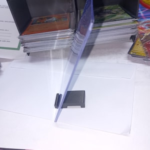 May include: A clear, protective card sleeve is held by a small, dark gray stand. Stacks of trading cards are visible in the background, organized in black storage containers. A white envelope is in the foreground.