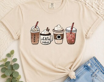 May include: A beige short-sleeve t-shirt featuring a graphic print of four beverage containers. The design includes a coffee cup with paw prints, a mug labeled CAT MOM, a whipped cream topped drink, and a cup filled with small pink heart shapes.
