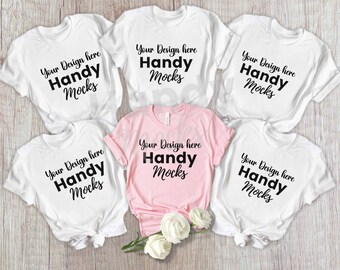 May include: Six t-shirts, including one pink and five white, are arranged on a light wood surface. Each shirt features the text "Your Design here Handy Mocks" in a bold, black font. The pink shirt is folded, and the white shirts are knotted.