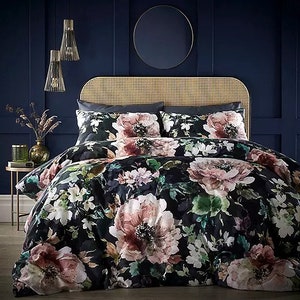 Puede incluir: Una funda nórdica negra con un estampado floral grande en tonos rosa, blanco y verde. La funda nórdica está en una cama con una cabecera tejida y dos fundas de almohada a juego.
