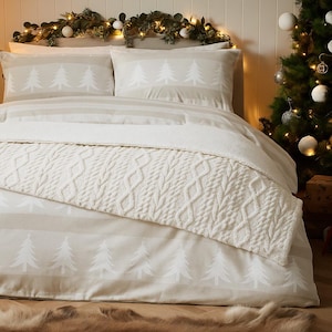 Op de afbeelding: Een beige en witte kerstbeddengoedset. De set bevat een dekbedovertrek en kussenslopen met een herhalend patroon van witte kerstbomen. Een crèmekleurige kabelgebreide sprei is over het bed gedrapeerd. Het bed is versierd met een slinger en een kerstboom.