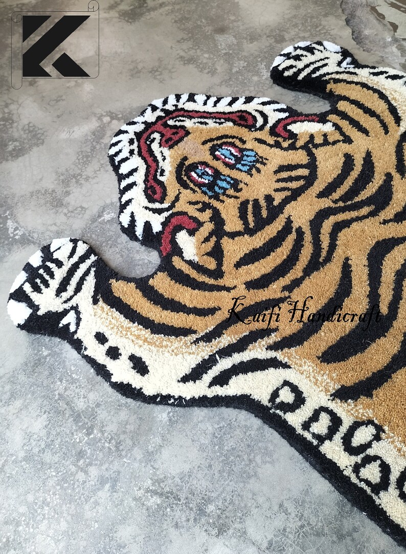 K&ouml;nnte beinhalten: Ein brauner und schwarzer Tigerteppich mit wei&szlig;en Streifen. Der Teppich ist aus flauschigem Garn gefertigt und hat einen cartoonartigen Stil. Die Augen des Tigers sind blau und der Mund ist in einem Br&uuml;llen ge&ouml;ffnet. Der Teppich ist mit "Kufti Handicraft" signiert.