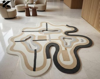 May include: A modern, abstract area rug with a unique, organic shape. The rug features a cream-colored base with flowing lines in shades of gray, black, and tan. The text "Kaifl Handioraft" is visible on the rug.