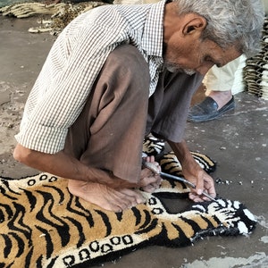 K&ouml;nnte beinhalten: Eine Person n&auml;ht von Hand einen Tiger-f&ouml;rmigen Teppich mit Nadel und Faden. Der Teppich besteht aus einem weichen, pl&uuml;schigen Material und zeigt ein realistisches Tigerdesign mit schwarzen Streifen und orangefarbenem Fell. Der Teppich liegt auf einem Betonboden.
