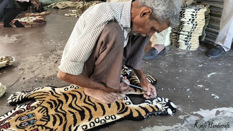 K&ouml;nnte beinhalten: Eine Person n&auml;ht von Hand einen Tiger-f&ouml;rmigen Teppich mit Nadel und Faden. Der Teppich besteht aus einem weichen, pl&uuml;schigen Material und zeigt ein realistisches Tigerdesign mit schwarzen Streifen und orangefarbenem Fell. Der Teppich liegt auf einem Betonboden.