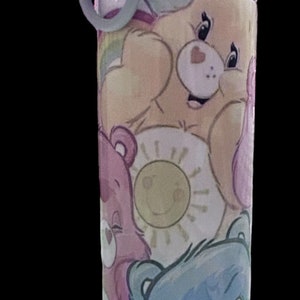 May include: A stainless steel water bottle with a gray lid and a carrying handle. The bottle features a colorful design of Care Bears characters, including a yellow bear with a heart on its chest, a pink bear with a sun, and a blue bear with a heart on its nose.