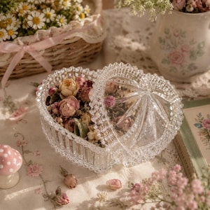 Puede incluir: Un joyero de cristal transparente en forma de corazón con un patrón de facetas talladas en diamante y una tapa extraíble. El recipiente está lleno de un surtido de flores secas en tonos rosa, crema y verde apagado, visibles a través del cristal.