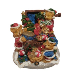 May include: A decorative figurine featuring three brown teddy bears in winter attire surrounding a wooden chest filled with Christmas gifts, a small green tree, and a drum, all set on a circular base with a white textured trim.
