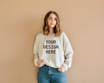 May include: A woman wearing a light beige sweatshirt with the text "YOUR DESIGN HERE" printed on the front. She is wearing blue jeans and standing in front of a light brown wall.