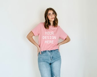 Puede incluir: Una camiseta rosa con el texto "YOUR DESIGN HERE" impreso en la parte delantera. La camiseta la lleva puesta una persona con el pelo largo y castaño y va combinada con unos vaqueros azules.