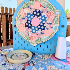 May include: A wooden embroidery hoop holds a patchwork of fabric hexagons in pink, blue, and green floral and polka dot patterns. The hoop is mounted on a light blue pegboard, accompanied by a small bowl of clear clips and a spool of white thread.