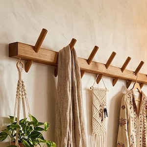 Op de afbeelding: Houten wandkapstok met schuine pinnen. Een beige sjaal, een macramé wandkleed en een jas met patroon hangen aan de pinnen. Een plant in een macramé hanger voegt groen toe.