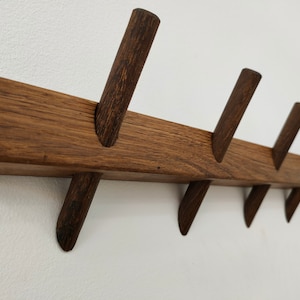 Könnte beinhalten: Ein Wand-Kleiderhaken aus Holz mit acht Haken. Der Haken ist aus dunkelbraunem Holz und hat eine natürliche Oberfläche. Die Haken sind abgewinkelt und ebenfalls aus dunkelbraunem Holz.